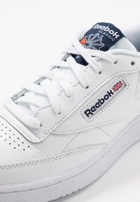 Basket Reebok Classic en cuir blanc avec lacets blancs, étiquette de languette bleu marine et petit drapeau Union Jack sur le patch du logo latéral.