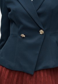 Blazer bleu marine en tissu texturé, à double boutonnage, avec des boutons dorés en forme de tête de lion, assorti à une jupe plissée de couleur rouille.