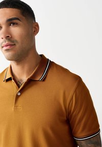 Polo tričko s krátkym rukávom v horčicovej žltej farbe s textúrovanou tkaninou. Má čierno-biely pásikavý golier a zapínanie na jeden gombík.