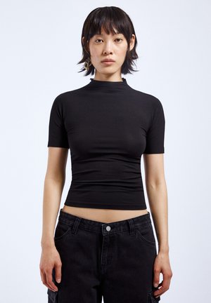 Black, fitted short-sleeve top with a high collar, made of smooth, stretchy material, paired with high-waisted black jeans.