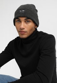 Gray beanie with a ribbed texture and a small black label featuring the Puma logo. Worn over a black turtleneck shirt.
