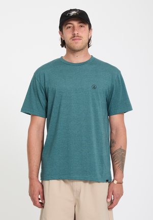 Homme portant un t-shirt à manches courtes bleu sarcelle, un short beige et une casquette noire, debout devant un fond blanc uni.