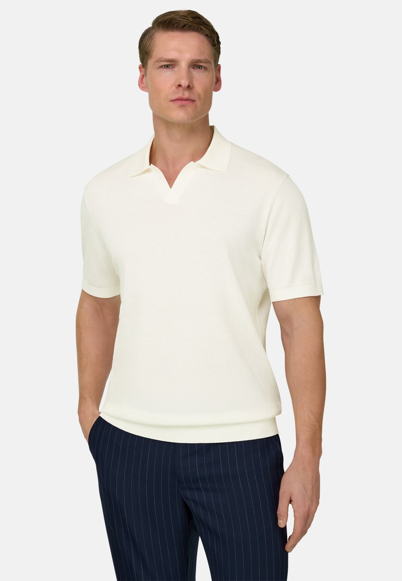 Homme portant un polo à manches courtes de couleur crème et un pantalon bleu marine à fines rayures, debout avec une main dans la poche, sur un fond uni.