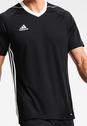 Man draagt een zwart Adidas sportshirt met witte strepen op de mouwen en een witte V-hals, staande tegen een lichte achtergrond.