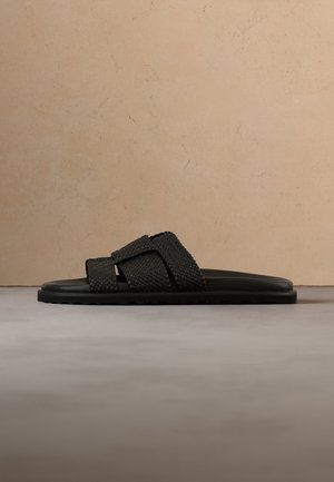 Mustad libisev sandaal tekstureeritud risti rihmadega ja lameda taldaga neutraalsel põrandal ning beežil taustal.