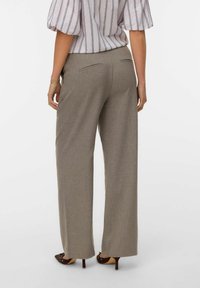 Donna che indossa pantaloni grigi a gamba larga e una camicetta a righe, in piedi con le mani rilassate lungo i fianchi, con i tacchi marroni visibili.