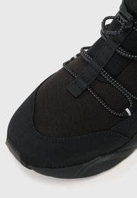 Μαύρο αθλητικό παπούτσι με υφασμάτινο άνω μέρος με υφή, προστατευτική μύτη και ελαστικά κορδόνια με ανακλαστικές λεπτομέρειες για αυξη�μένη ορατότητα.