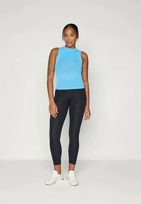 Lys blå ribbet ermeløs topp kombinert med svarte leggings og hvite sneakers. Modellen står med hendene på hoftene mot en ensfarget bakgrunn.