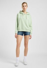Sudadera con capucha de color verde claro con cordones blancos, combinada con shorts de mezclilla y zapatillas blancas. Presenta un logo sutil en el pecho.