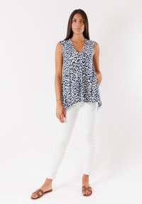 Top senza maniche in tessuto bianco con stampa leopardata blu navy; design a V, vestibilità morbida, dotato di tasche laterali, abbinato a pantaloni bianchi e sandali marroni.