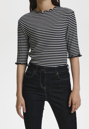 Frau trägt ein schwarz-weiß gestreiftes, geripptes Shirt mit wellenförmigen Kanten und hoch taillierte dunkelblaue Jeans.