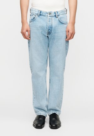 Personne portant un jean ample bleu clair et des chaussures habillées noires, debout, les bras détendus le long du corps, devant un fond uni.