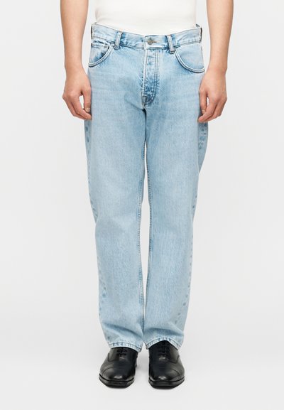 Personne portant un jean ample bleu clair et des chaussures habillées noires, debout, les bras détendus le long du corps, devant un fond uni.