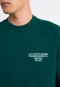 Mørkegrøn sweatshirt med ribbet rund hals, featuring hvid broderet "CARHARTT WIP" logo på venstre brystområde. Blød tekstur.