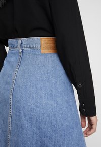 Jeanskjol med hög midja, ljusblått tyg och en brun läderetikett med varumärkesdetaljer. Knäppt svart långärmad skjorta synlig.