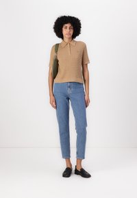 Polo shirt beige lavorato a maglia con colletto, abbinato a jeans dritti azzurri chiari e mocassini neri. Il modello porta una borsa a tracolla verde.