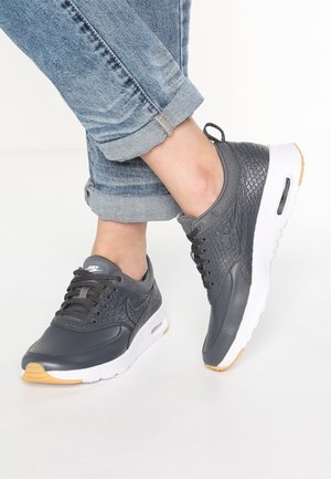 Personne portant des baskets Nike gris foncé texturées avec des semelles blanches et un jean bleu clair retroussé, debout sur un sol blanc.