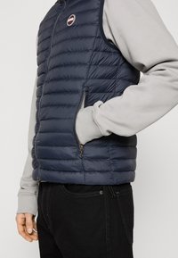 Navy puffer vest with horizontal quilting, side pockets, and a round logo patch. Worn over a gray sweatshirt with cuffed sleeves.