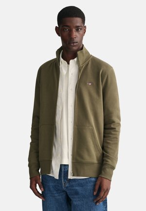 Sudadera de cremallera en verde oliva con cuello alzado, dos bolsillos frontales, puños acanalados y un pequeño logo en el lado izquierdo del pecho.