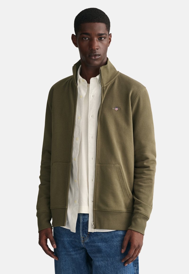 Sudadera de cremallera en verde oliva con cuello alzado, dos bolsillos frontales, puños acanalados y un pequeño logo en el lado izquierdo del pecho.