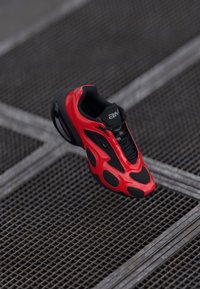 Zapatilla atlética roja con un diseño aerodin�ámico, que presenta acentos de malla negra, cordones visibles y una suela de goma, situada sobre un fondo texturizado.