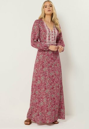 Femme portant une robe longue à manches longues, rose à motifs floraux, avec un décolleté en V et des panneaux blancs à motifs, debout les mains jointes devant elle.