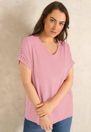 Kobieta w różowej bluzce z krótkim rękawem i dekoltem w kształcie litery V z okrągłymi wycięciami na rękawach, pozująca z rękami skrzyżowanymi na tle jednolitego beżowego tła.