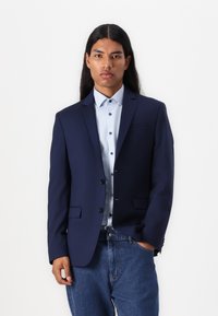 Navy blazer with a fitted design, notched lapels, and two front pockets, paired with a light blue button-up shirt and denim jeans.