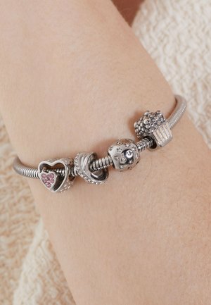 Silver charm bracelet featuring a heart with pink stones, a seashell, a star-patterned bead, and a textured cupcake charm on a flexible chain.