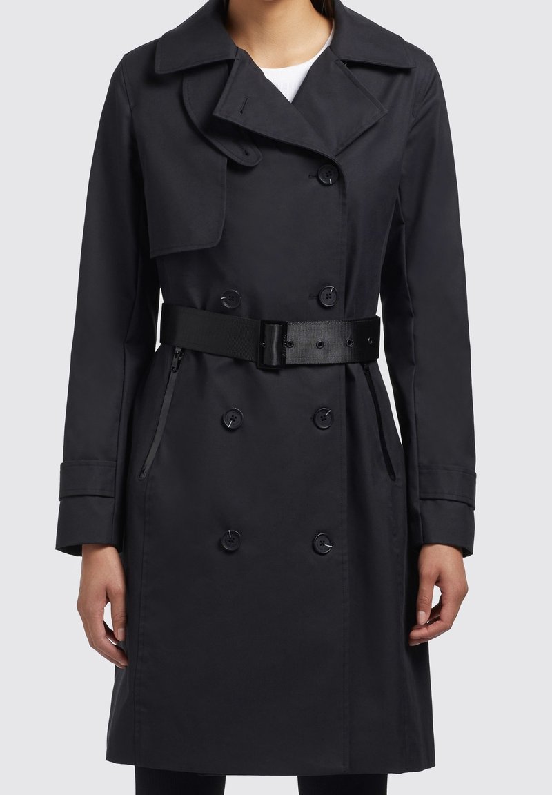 Trench-coat noir croisé avec un large col, une taille ceinturée et des poches zippées à l'avant. Caractéristiques de tissu lisse et de grands boutons.