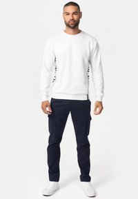 Sudadera blanca con diseño de franja negra lateral, combinada con pantalones cargo oscuros en azul marino y zapatillas bajas blancas. Outfit casual.