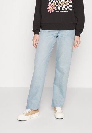 Partie inférieure du corps d'une personne portant un jean bleu clair, des baskets beige et blanches, et un sweat-shirt noir avec un texte coloré et des motifs floraux.