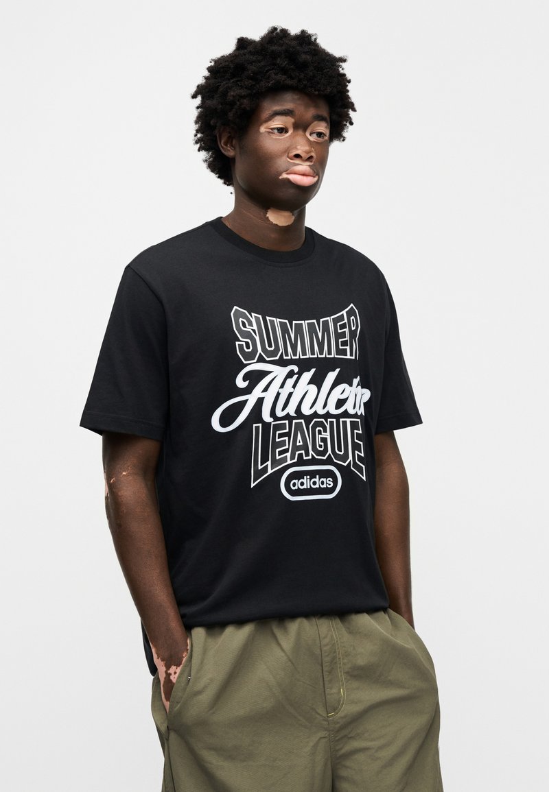 Młody mężczyzna z bielactwem, ubrany w czarną koszulkę Adidas z napisem „Summer Athlete League” oraz oliwkowe spodnie, stojący z rękami w kieszeniach.