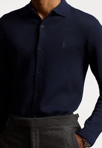 Marinblus med långa ärmar i texturerad tyg, med knäppt krage, svarta knappar och en liten broderad logotyp på bröstet.