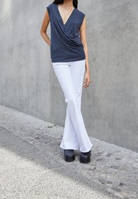 Graues, drapiertes Oberteil mit tiefem V-Ausschnitt, kombiniert mit weißen, ausgestellten Hosen und schwarzen Plateauschuhen, vor einem Betonhintergrund.