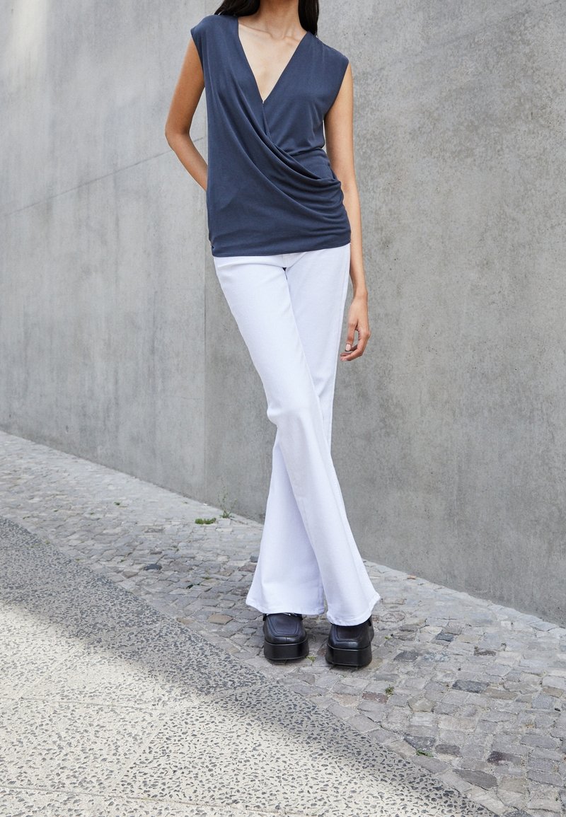 Graues, drapiertes Oberteil mit tiefem V-Ausschnitt, kombiniert mit weißen, ausgestellten Hosen und schwarzen Plateauschuhen, vor einem Betonhintergrund.