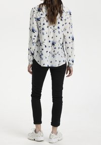 Chemisier blanc à imprimé floral bleu, à manches longues et avec un bouton sur le devant. Associé à un pantalon noir ajusté et des baskets claires.