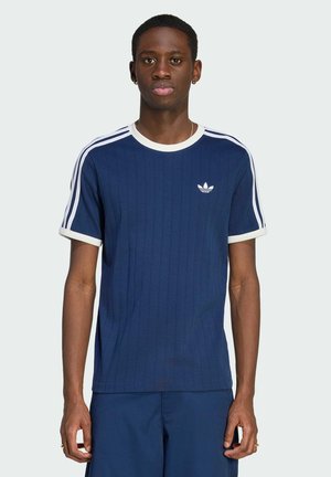Mladý muž nosící tmavě modré tričko Adidas s bílými pruhy na rukávech a logem na hrudi, stojící před jednobarevným světlým pozadím.