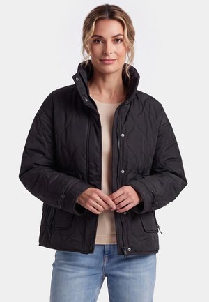 Donna che indossa una giacca nera trapuntata sopra una camicia beige e jeans azzurri chiari, in piedi davanti a uno sfondo chiaro e semplice.
