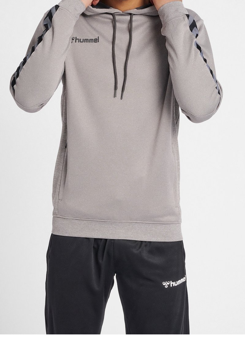 Sudadera gris con un diseño texturizado, cordones y acentos negros en los laterales. Combinada con pantalones negros que tienen un logo blanco en la pierna.