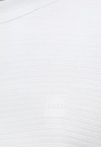 Balts teksturēts audums ar horizontālām ribām, apakšējā labajā stūrī ar nelielu baltu kvadrātveida etiķeti, uz kuras ir izspiests uzraksts "BOSS".