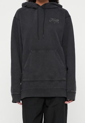 Osoba ubrana w za dużą, wyblakłą czarną bluzę z kapturem z przednią kieszenią i wyhaftowanym napisem „Hugo” na lewej piersi, w połączeniu z luźnymi czarnymi spodniami.