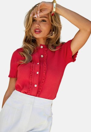 Žena s dlhými blond vlasmi, oblečená v červenom krátkom tričku s volánmi a bielymi gombíkmi, v kombinácii s bielymi nohavicami s vysokým pásom, si rukou zakrýva oči.