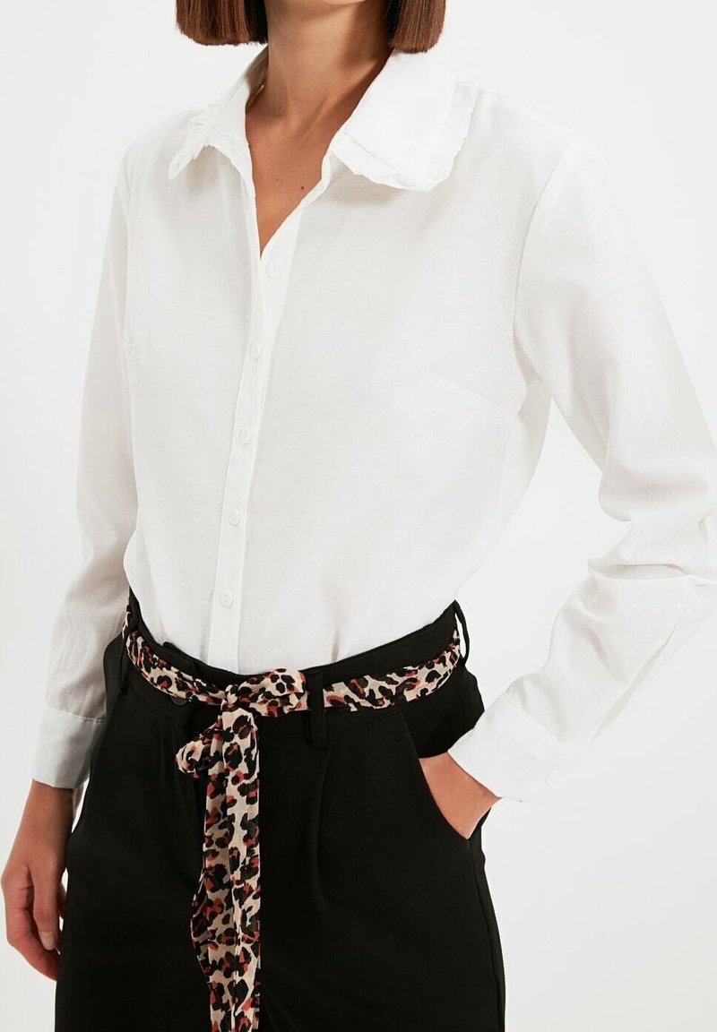 Femme portant un chemisier blanc à boutons avec un col à volants et un pantalon noir ceinturé d'une ceinture en tissu imprimé léopard.