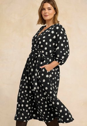 Frau trägt ein schwarzes Midikleid mit weißen Polka-Dots, dreiviertel Ärmeln, einem Wickelgürtel in der Taille und hat die Hände in den Taschen des Kleides.