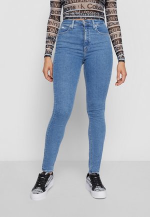 Personne portant un jean skinny bleu clair, un haut en mesh avec le logo Calvin Klein, et des baskets plateformes noires et blanches avec impression de texte.