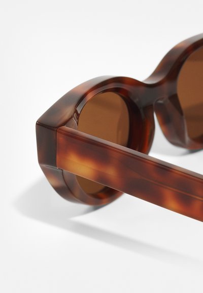 Ochelari de soare maro cu model broască țestoasă, cu un cadru geometric voluminos și lentile maro închise. Textura netedă și brațele groase îmbunătățesc durabilitatea.