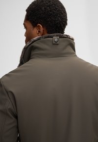 Ein Mann von hinten gesehen, der eine braune Jacke mit einem hochgeschlagenen Kragen trägt, der mit weichem Fell ausgekleidet ist, vor einem einfarbigen hellen Hintergrund.