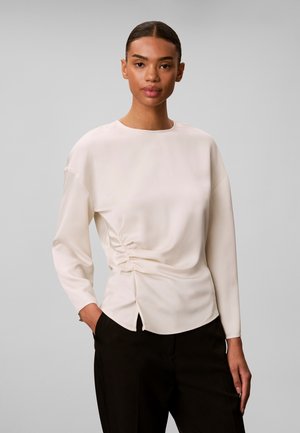 Jeune femme aux cheveux foncés portant un haut beige clair à manches longues avec fronces latérales et un pantalon noir, debout devant un fond gris uni.