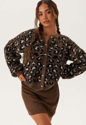 Kvinna med flätat hår som bär en brun leopardmönstrad fluffig cardigan med pärlknappar och en matchande brun kjol, poserar med händerna på höfterna.